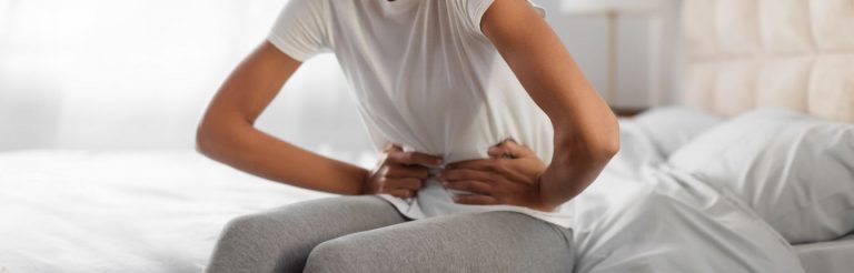 A close up of a woman sitting on a bed, holding her stomach as if experiencing cramps.