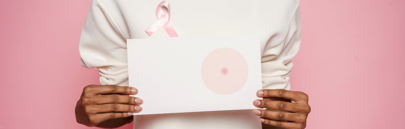 A woman in a white sweater holding up a piece of paper showing a drawing of a breast. She is also wearing a breast cancer ribbon.