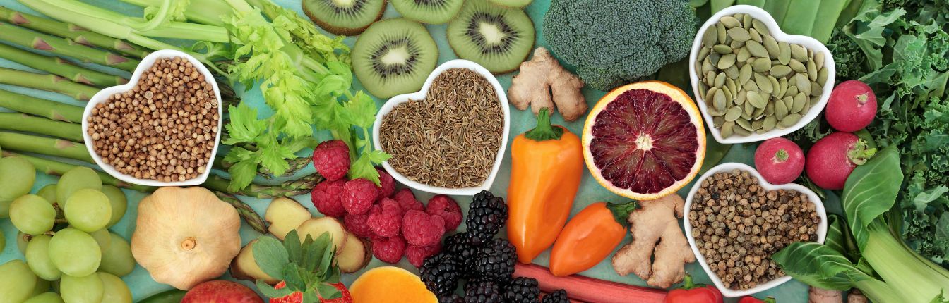 A variety of fruits and vegetables, with small white dishes in the shape of hearts holding nuts and seeds.